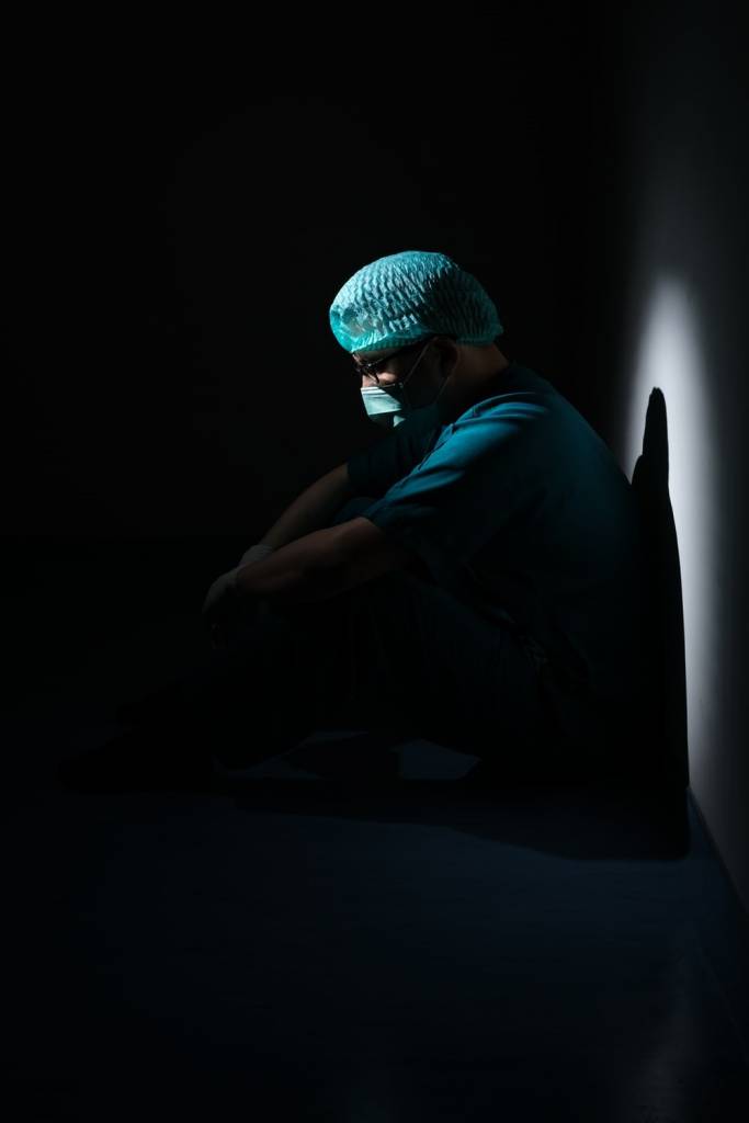 A Partially Visible Nurse in Scrubs and Mask Sitting by a Wall, and Looking Down While Looking Visibly Depressed